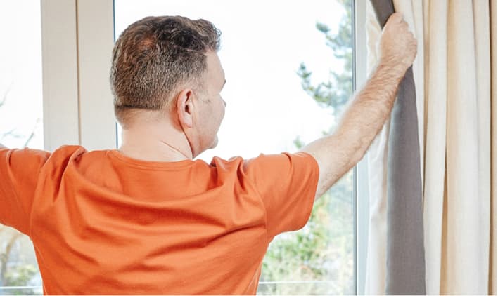 A man opening curtains and looking out a window.